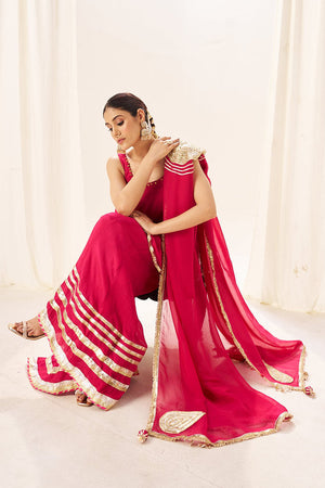 Woman in a pink sharara set and orgenza dupatta with gold accents sitting against a white curtain.